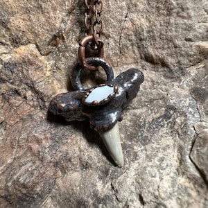 May include: A handcrafted copper pendant necklace featuring a fossilized shark tooth. The pendant has a dark, textured finish with a small, light-colored stone accent. The chain is made of copper-colored links.
