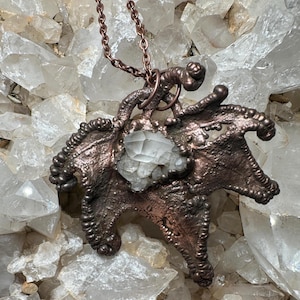 May include: A handcrafted copper pendant necklace featuring a unique, textured design with a central, clear quartz crystal. The pendant is suspended from a delicate copper chain. The necklace is displayed against a backdrop of clear quartz crystals.