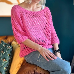 Peut inclure: Un pull en crochet rose à la coupe ample et au motif ajouré délicat. Le pull a un col large et des manches trois-quarts.