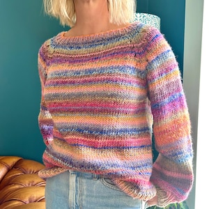 Könnte beinhalten: Ein handgestrickter Pullover mit Bootsausschnitt und langen Ärmeln. Der Pullover hat horizontale Streifen in verschiedenen Farben, darunter Rosa, Orange, Blau und Lila. Das Kleidungsstück ist aus einem dicken, strukturierten Garn gefertigt.