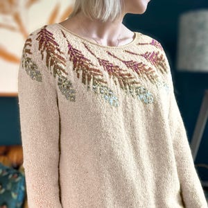 Könnte beinhalten: Ein cremefarbener Strickpullover mit einem bunten Blattmotiv am Ausschnitt.