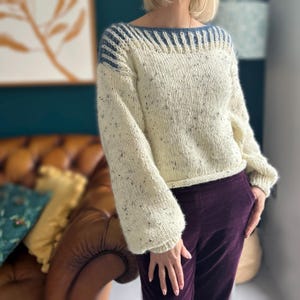 May include: A white knitted jumper with a blue and white striped neckline. The jumper has a speckled texture and a cropped length.