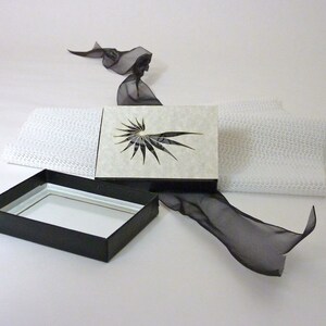 以下が含まれることがあります： 白の蓋が付いた黒い箱。蓋が開いており、黒と白の幾何学模様が見える。箱は白い表面に置かれ、黒いリボンが置かれている。
