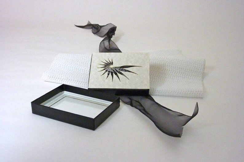 以下が含まれることがあります： 白の蓋が付いた黒い箱。蓋が開いており、黒と白の幾何学模様が見える。箱は白い表面に置かれ、黒いリボンが置かれている。