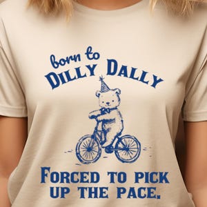 May include: A beige t-shirt with a blue graphic of a bear wearing a party hat riding a bicycle. The text "born to dilly dally" is above the bear and the text "forced to pick up the pace" is below the bear.