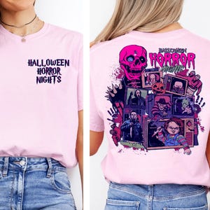 May include: Light pink t-shirt with the text "HALLOWEEN HORROR NIGHTS" on the front and a graphic on the back featuring a pink skull and various horror movie characters. The shirt is paired with blue denim jeans.
