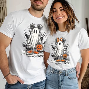 May include: White t-shirts featuring a Halloween-themed design. The graphic depicts a ghost wearing Mickey Mouse ears and holding a jack-o'-lantern bucket. The ghost is wearing orange shoes. The design is set against a black splatter background.
