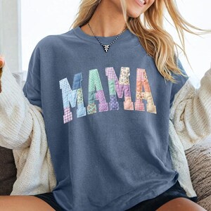May include: A blue-grey t-shirt with the word "MAMA" in patchwork letters. The letters are made of various floral and patterned fabrics. The shirt is worn with a white cardigan and black shorts.