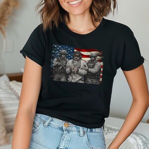 Pode incluir: T-shirt preto com um gr&aacute;fico de tr&ecirc;s soldados em frente a uma bandeira americana desgastada. Os soldados s&atilde;o retratados em uniforme militar, segurando armas. O design da bandeira incorpora as cores vermelha, branca e azul.