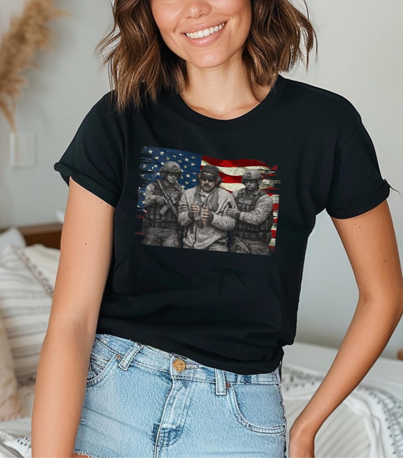 Pode incluir: T-shirt preto com um gr&aacute;fico de tr&ecirc;s soldados em frente a uma bandeira americana desgastada. Os soldados s&atilde;o retratados em uniforme militar, segurando armas. O design da bandeira incorpora as cores vermelha, branca e azul.