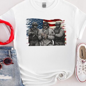Pode incluir: Camiseta branca com um gr&aacute;fico de tr&ecirc;s soldados em frente a uma bandeira americana desgastada. Os soldados usam equipamento militar. A camisa tem gola redonda e mangas curtas. A marca "GILDAN" &eacute; vis&iacute;vel na etiqueta.