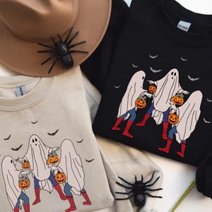 May include: Two sweatshirts, one beige and one black, featuring a Halloween-themed graphic. The design shows three ghosts carrying jack-o'-lanterns, with bats flying above. A brown hat and a black spider are also in the image.