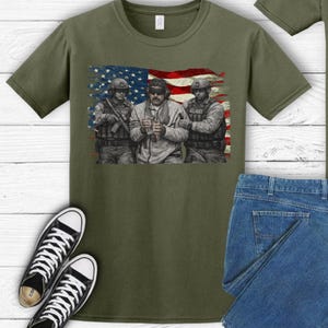 Pode incluir: Camiseta verde-oliva com um gr&aacute;fico de tr&ecirc;s soldados em frente a uma bandeira americana. A camisa &eacute; combinada com jeans azuis e t&ecirc;nis de lona pretos e brancos.
