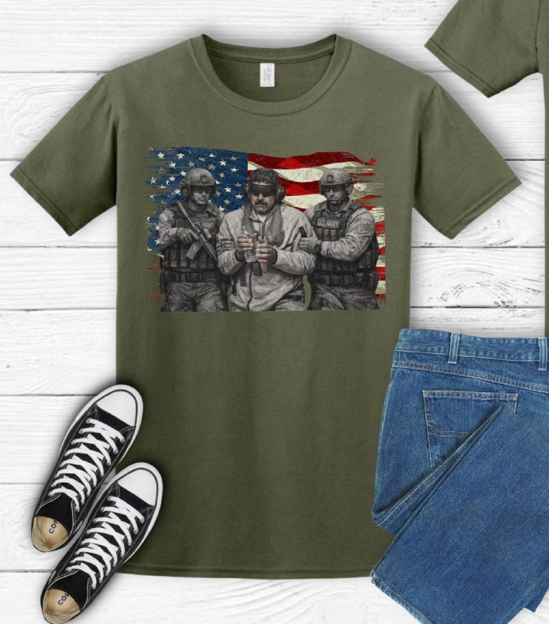 Pode incluir: Camiseta verde-oliva com um gr&aacute;fico de tr&ecirc;s soldados em frente a uma bandeira americana. A camisa &eacute; combinada com jeans azuis e t&ecirc;nis de lona pretos e brancos.