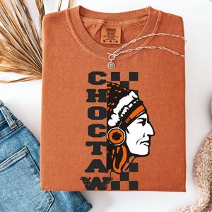 K&ouml;nnte beinhalten: Ein rostfarbenes T-Shirt mit dem Wort "CHOCTAW" vertikal in schwarzen Blockbuchstaben gestapelt. Eine Grafik eines indianischen Mannes im Profil befindet sich auf der rechten Seite. Eine silberne Halskette mit einem Sonnenstrahl-Anh&auml;nger liegt &uuml;ber dem Shirt.