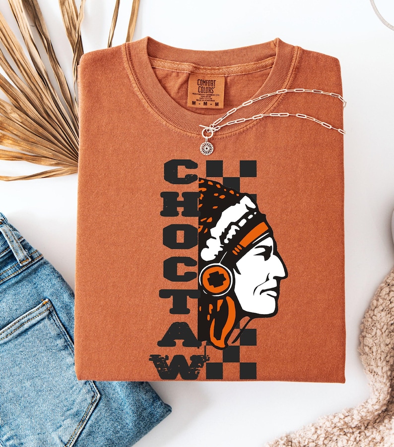 K&ouml;nnte beinhalten: Ein rostfarbenes T-Shirt mit dem Wort "CHOCTAW" vertikal in schwarzen Blockbuchstaben gestapelt. Eine Grafik eines indianischen Mannes im Profil befindet sich auf der rechten Seite. Eine silberne Halskette mit einem Sonnenstrahl-Anh&auml;nger liegt &uuml;ber dem Shirt.