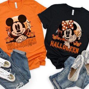 May include: Two Halloween-themed t-shirts, one orange and one black, featuring Mickey and Minnie Mouse designs. The shirts have the word "HALLOWEEN" printed in orange. The shirts are paired with blue jeans and shoes.