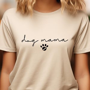 K&ouml;nnte beinhalten: Ein beiges T-Shirt mit dem Text "dog mama" in schwarzer Schrift und einem Pfotenabdruck mit einem Knochen darin.