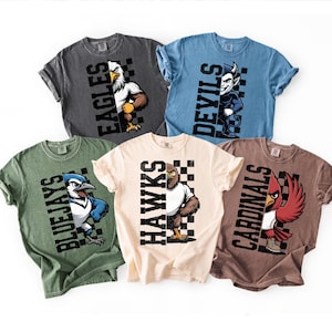 K&ouml;nnte beinhalten: F&uuml;nf kurz&auml;rmelige T-Shirts in verschiedenen Farben, jedes mit einem Teammaskottchen und -namen. Die Shirts sind grau, blau, gr&uuml;n, cremefarben und braun. Zu den Maskottchen geh&ouml;ren ein Adler, ein Teufel, ein Blue Jay, ein Hawk und ein Kardinal.