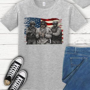 Pode incluir: Uma camiseta cinza claro com um gr&aacute;fico de tr&ecirc;s soldados em frente a uma bandeira americana. Os soldados est&atilde;o usando equipamento militar. A imagem &eacute; impressa em uma camiseta cinza claro, com um par de t&ecirc;nis preto e branco e jeans.