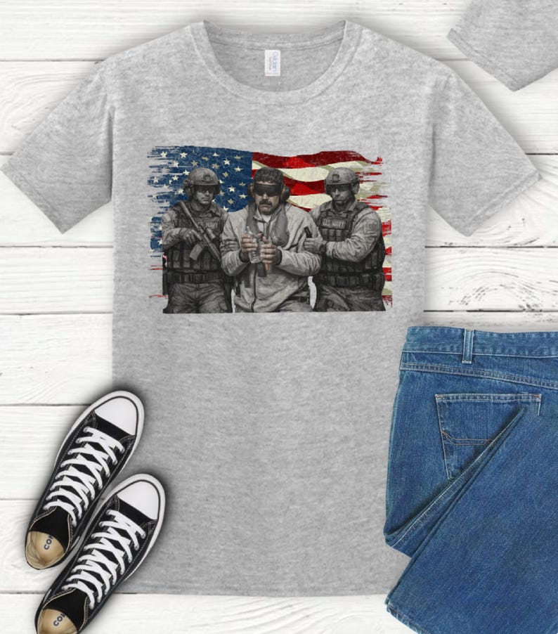 Pode incluir: Uma camiseta cinza claro com um gr&aacute;fico de tr&ecirc;s soldados em frente a uma bandeira americana. Os soldados est&atilde;o usando equipamento militar. A imagem &eacute; impressa em uma camiseta cinza claro, com um par de t&ecirc;nis preto e branco e jeans.