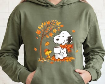 May include: Olive green hoodie featuring a Snoopy and Woodstock graphic. Snoopy is seated, wearing an orange scarf, and holding a white mug. Woodstock is standing nearby. The graphic includes a tree with orange leaves and fallen leaves.