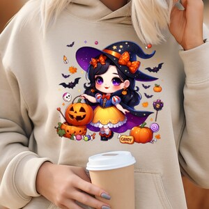 May include: Beige hoodie featuring a Halloween-themed graphic. The graphic depicts a cartoon witch with a pumpkin, bats, and the word "CANDY". The witch is wearing a purple hat with an orange bow and a yellow dress.