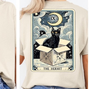 May include: Beige t-shirt featuring a tarot card design. The graphic depicts a black cat in a box under a crescent moon and stars, with the text "THE HERMIT" at the bottom. The card has a vintage, illustrated style.