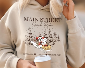 May include: Beige hoodie featuring a graphic of Mickey and Minnie Mouse on a sleigh with the text "Main Street Sleigh Rides". The design includes pine trees and the phrases "Hot Cocoa, Cookies, Magical Fun".