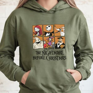 May include: Olive green hooded sweatshirt featuring a graphic of characters from "The Nightmare Before Christmas." The design includes six character portraits and the text "est.1993 THE NIGHTMARE BEFORE CHRISTMAS" in a black font. The sweatshirt has a front pocket.
