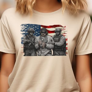 Pode incluir: Camiseta bege com um gr&aacute;fico de tr&ecirc;s soldados em frente a uma bandeira americana desgastada. Os soldados usam equipamento militar e seguram armas. A imagem tem uma est&eacute;tica vintage e patri&oacute;tica.