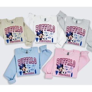 K&ouml;nnte beinhalten: Eine Auswahl an Rundhals-Sweatshirts in Beige, Wei&szlig;, Grau, Hellblau und Rosa. Jedes Sweatshirt zeigt das Wort "BUFFALO" in Burgunderrot, zusammen mit einer Cartoonfigur und Football-Grafiken.