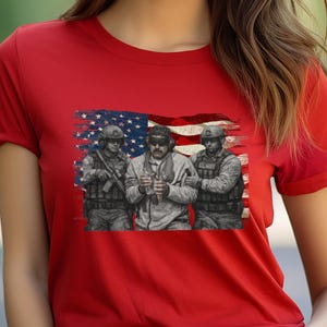 Pode incluir: Camiseta vermelha com uma estampa de tr&ecirc;s soldados em frente a uma bandeira americana desgastada. Os soldados usam equipamento militar. A estampa &eacute; em tons de cinza e preto.
