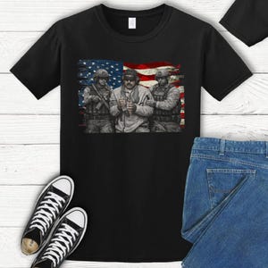 Pode incluir: Camiseta preta com um gr&aacute;fico de tr&ecirc;s soldados em frente a uma bandeira americana. Os soldados est&atilde;o usando equipamento militar. A imagem tamb&eacute;m inclui t&ecirc;nis preto e branco e jeans azul.