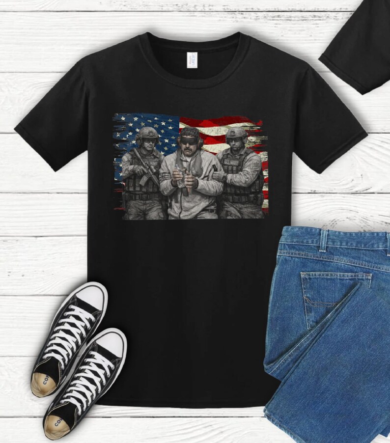 Pode incluir: Camiseta preta com um gr&aacute;fico de tr&ecirc;s soldados em frente a uma bandeira americana. Os soldados est&atilde;o usando equipamento militar. A imagem tamb&eacute;m inclui t&ecirc;nis preto e branco e jeans azul.