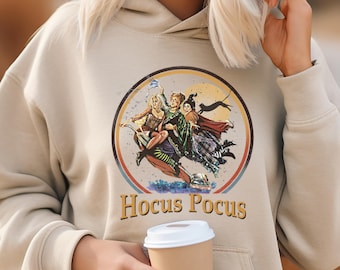 May include: Beige hoodie featuring a circular graphic with three witches riding a broom, a black cat, and the text "Hocus Pocus." The graphic is in a vintage style. The hoodie has a drawstring hood and a front pocket.