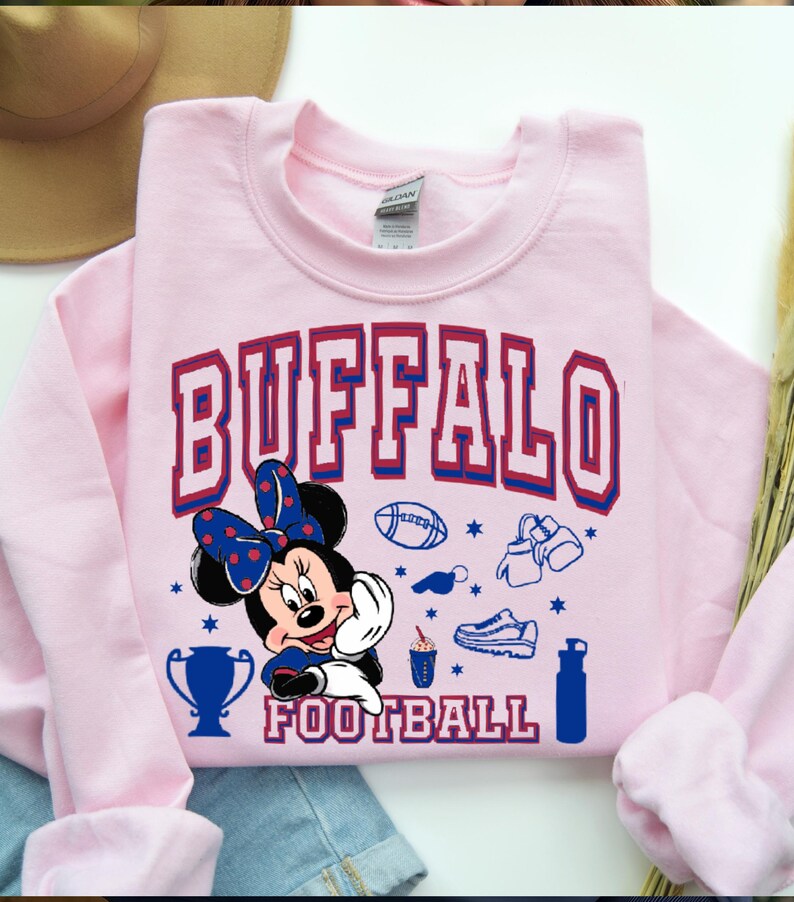 K&ouml;nnte beinhalten: Ein hellrosa Sweatshirt mit dem Wort "BUFFALO" in roten und blauen Blockbuchstaben. Eine Cartoon-Minnie-Maus mit einer blauen Schleife ist unter dem Wort "BUFFALO" abgebildet. Das Wort "FOOTBALL" steht darunter. Fu&szlig;ball-Grafiken sind ebenfalls enthalten.