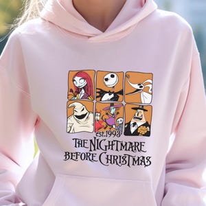 May include: Light pink hoodie featuring a graphic from "The Nightmare Before Christmas." The design includes character portraits in orange squares and the film's title. The text "est.1993" is also included.