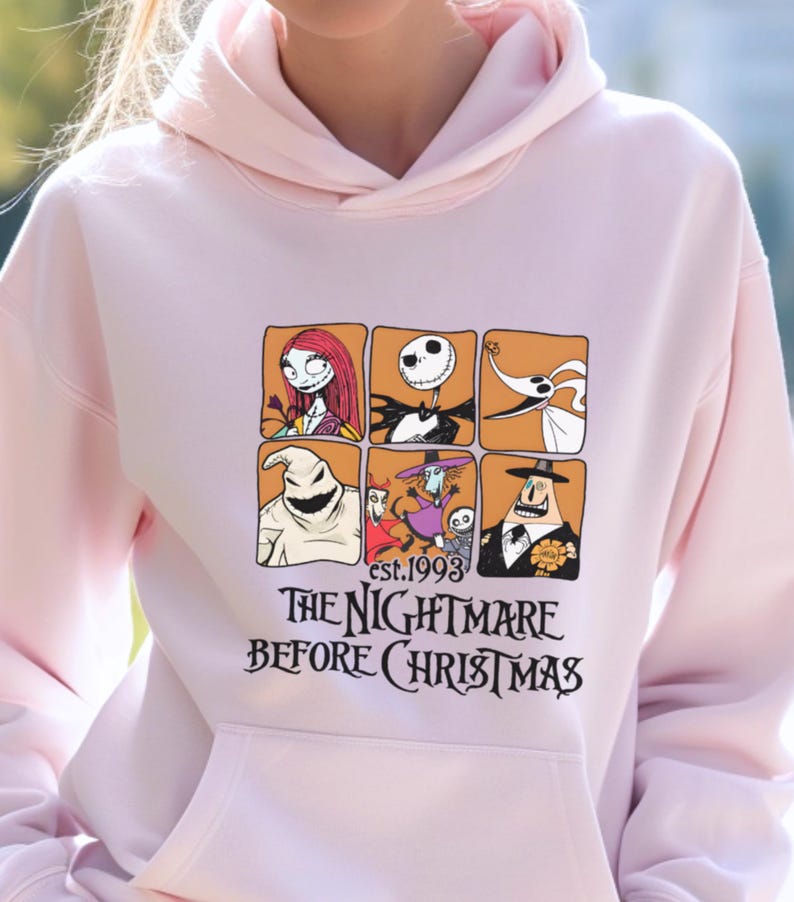 May include: Light pink hoodie featuring a graphic from "The Nightmare Before Christmas." The design includes character portraits in orange squares and the film's title. The text "est.1993" is also included.