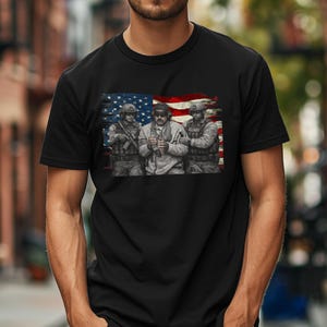 Pode incluir: T-shirt preto com um gr&aacute;fico de tr&ecirc;s soldados em uniforme militar, contra um fundo da bandeira americana. A imagem &eacute; uma ilustra&ccedil;&atilde;o detalhada com uma est&eacute;tica vintage e desgastada.