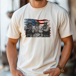 Pode incluir: T-shirt creme com um gr&aacute;fico de tr&ecirc;s soldados em frente a uma bandeira americana desgastada. Os soldados s&atilde;o retratados em tons de cinza, com a bandeira em vermelho, branco e azul. A t-shirt &eacute; feita de um material macio.