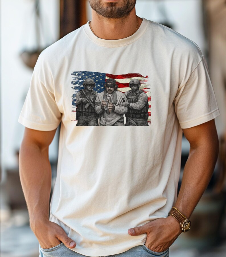 Pode incluir: T-shirt creme com um gr&aacute;fico de tr&ecirc;s soldados em frente a uma bandeira americana desgastada. Os soldados s&atilde;o retratados em tons de cinza, com a bandeira em vermelho, branco e azul. A t-shirt &eacute; feita de um material macio.