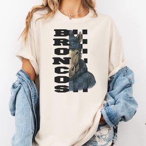K&ouml;nnte beinhalten: Cremefarbenes T-Shirt mit einer Pferdeillustration, die eine blaue M&uuml;tze und Jacke tr&auml;gt. Das Wort "BRONCOS" ist vertikal in einem schwarz-wei&szlig;en Schachbrettmuster gedruckt. Das Wort "VER" ist auf der M&uuml;tze aufgedruckt.