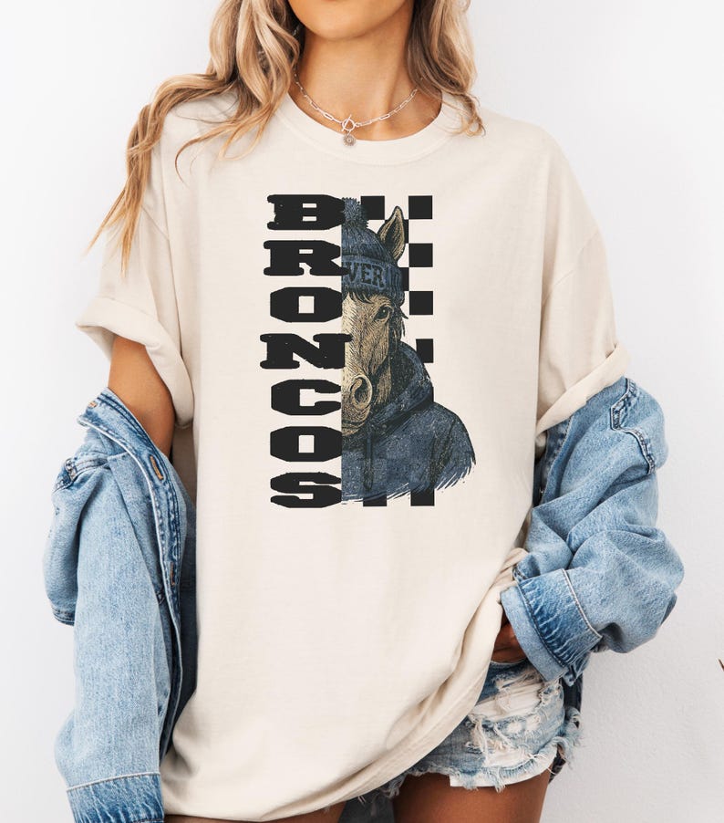 K&ouml;nnte beinhalten: Cremefarbenes T-Shirt mit einer Pferdeillustration, die eine blaue M&uuml;tze und Jacke tr&auml;gt. Das Wort "BRONCOS" ist vertikal in einem schwarz-wei&szlig;en Schachbrettmuster gedruckt. Das Wort "VER" ist auf der M&uuml;tze aufgedruckt.