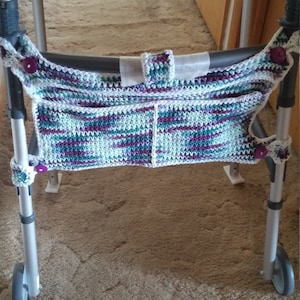 May include: A silver walker with a crocheted bag attached to the handles. The bag is white with purple, teal, and green stripes. The walker is on a beige carpet.