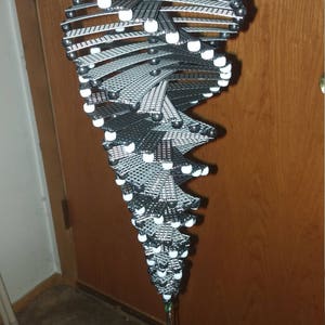May include: A black and white beaded mobile with a spiral design. The mobile is hanging from a gold hook and has a small black and white beaded figure at the bottom.