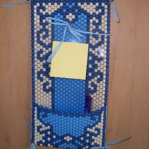May include: A blue and white beaded wall organizer with a pocket for holding pens or pencils. A yellow sticky note is attached to the organizer.