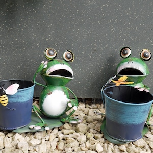 May include: Two green metal frog figurines, each featuring a wide-eyed expression and a large open mouth. Each frog is attached to a blue cylindrical metal planter bucket. One bucket features a yellow and black bee, while the other has a small orange butterfly.