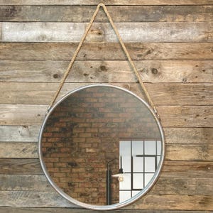 52cm diameter industrial style round mirror hanging on wall by its jute rope hanger, the frame is made from metal and finished in a rustic silver.