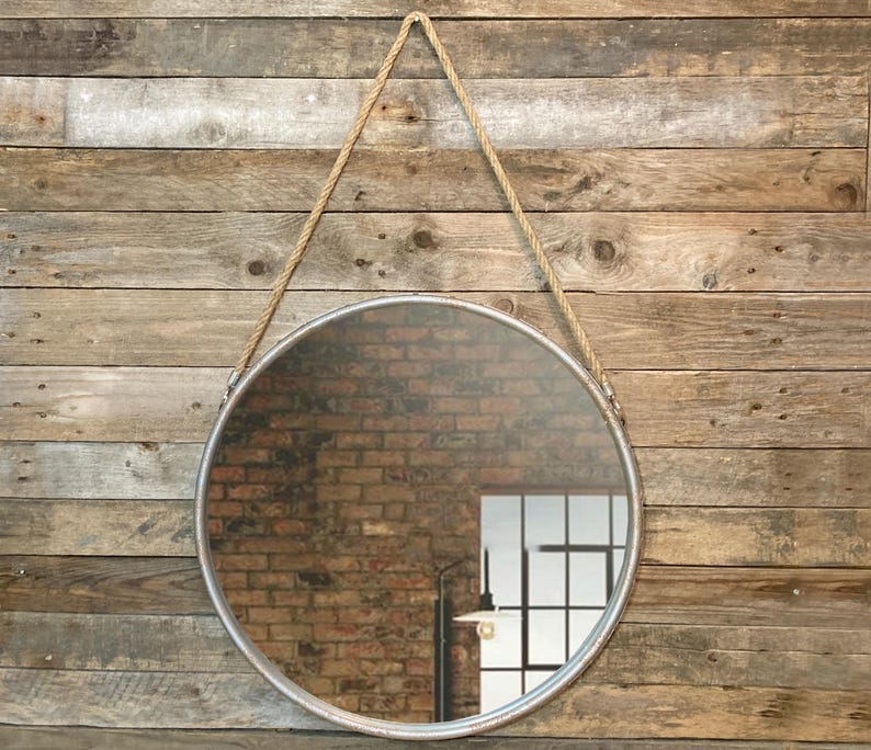 52cm diameter industrial style round mirror hanging on wall by its jute rope hanger, the frame is made from metal and finished in a rustic silver.